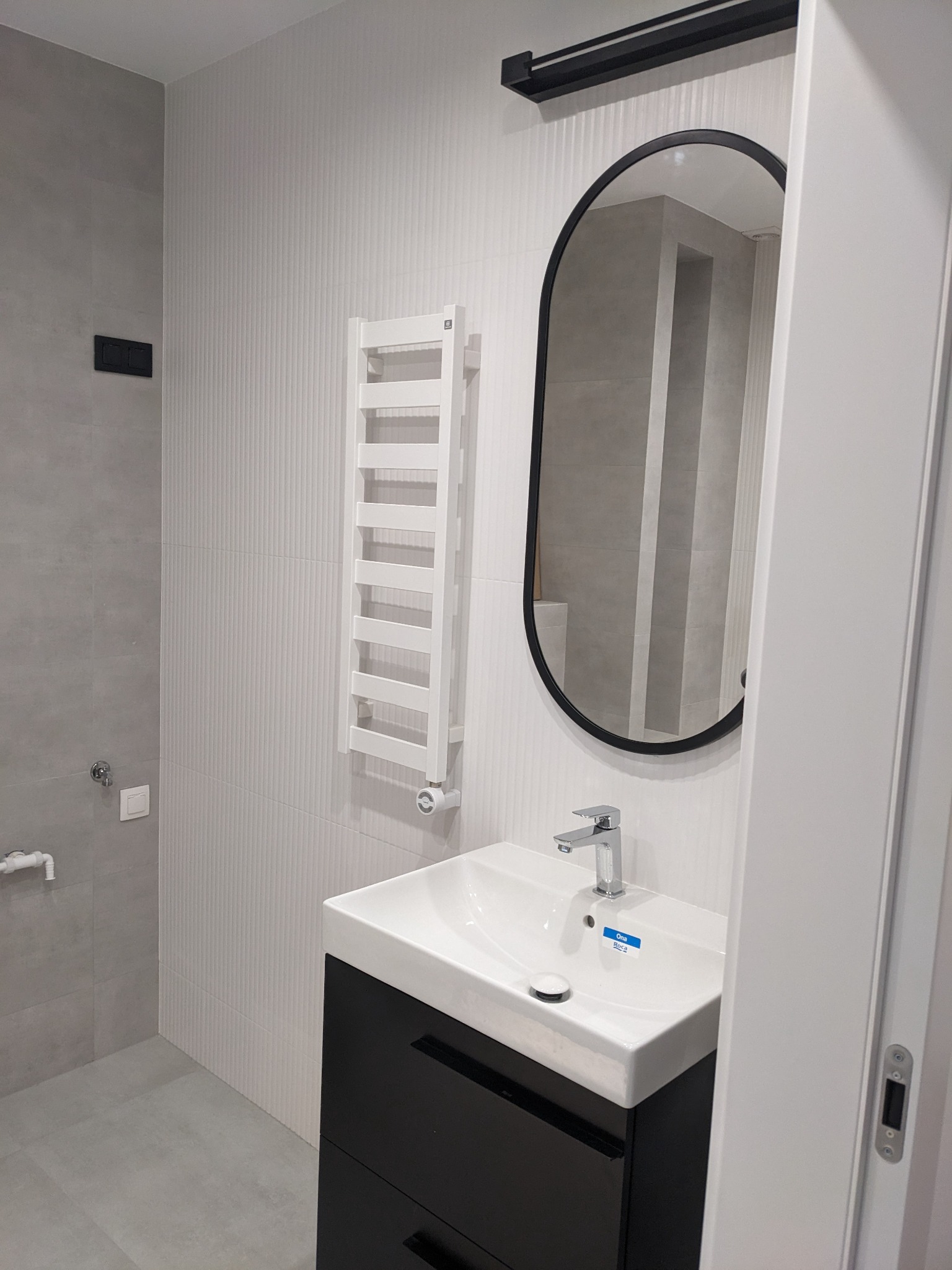 Nowoczesna łazienka w szaro-białej kolorystyce z owalnym lustrem, grzejnikiem drabinkowym i czarną szafką pod umywalką z baterią Roca Ona, widok zza uchylonych drzwi.