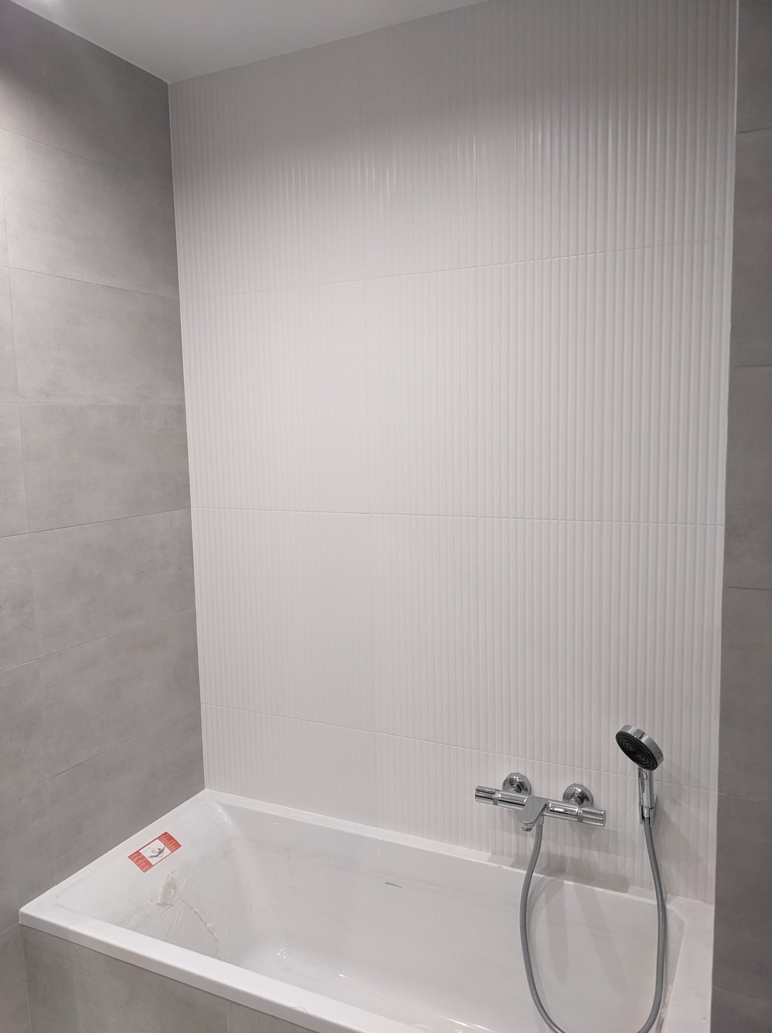 Biała wanna obudowana szarą płytką, z chromowaną baterią i słuchawką prysznicową na tle ściany wyłożonej pionowymi, białymi płytkami strukturalnymi.
