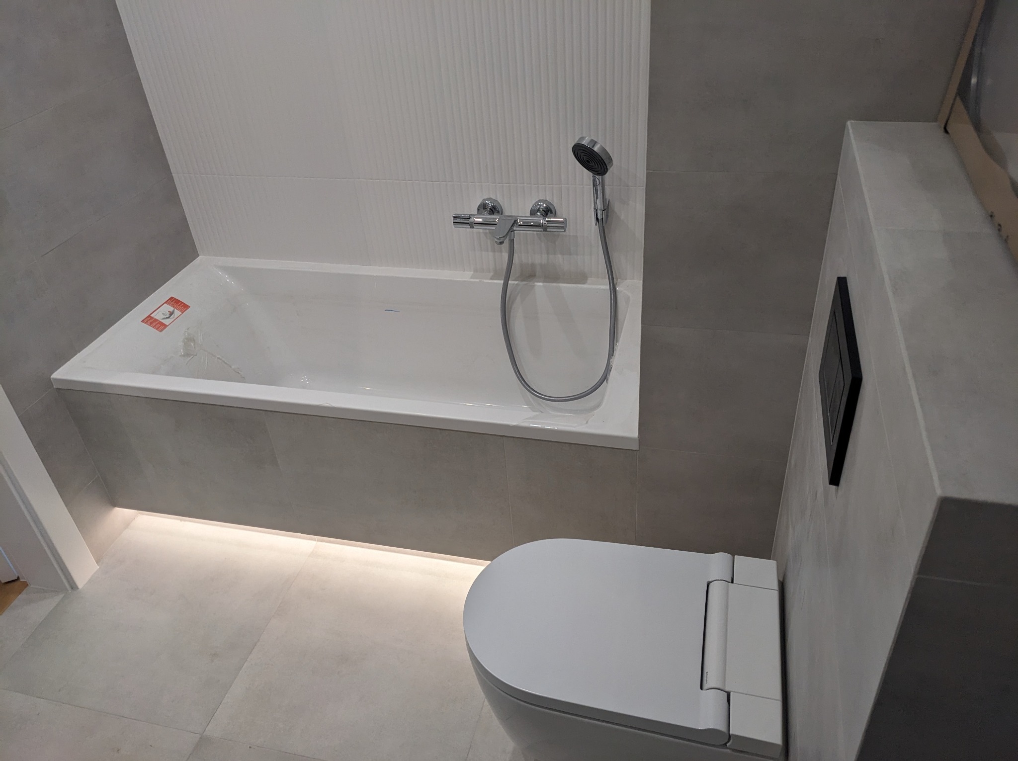 Nowoczesna łazienka z szarą płytką, białą wanną z ukrytym oświetleniem LED i wiszącą toaletą, minimalistyczny design.