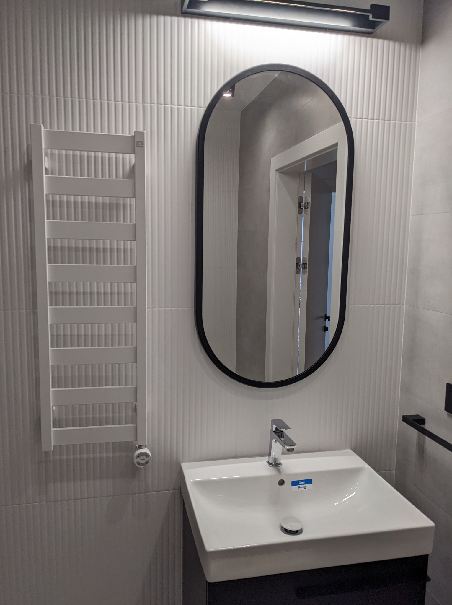 Nowoczesna łazienka z umywalką na szafce, owalnym lustrem w czarnej ramie i grzejnikiem drabinkowym na ścianie pokrytej białymi, pionowo żłobionymi płytkami.