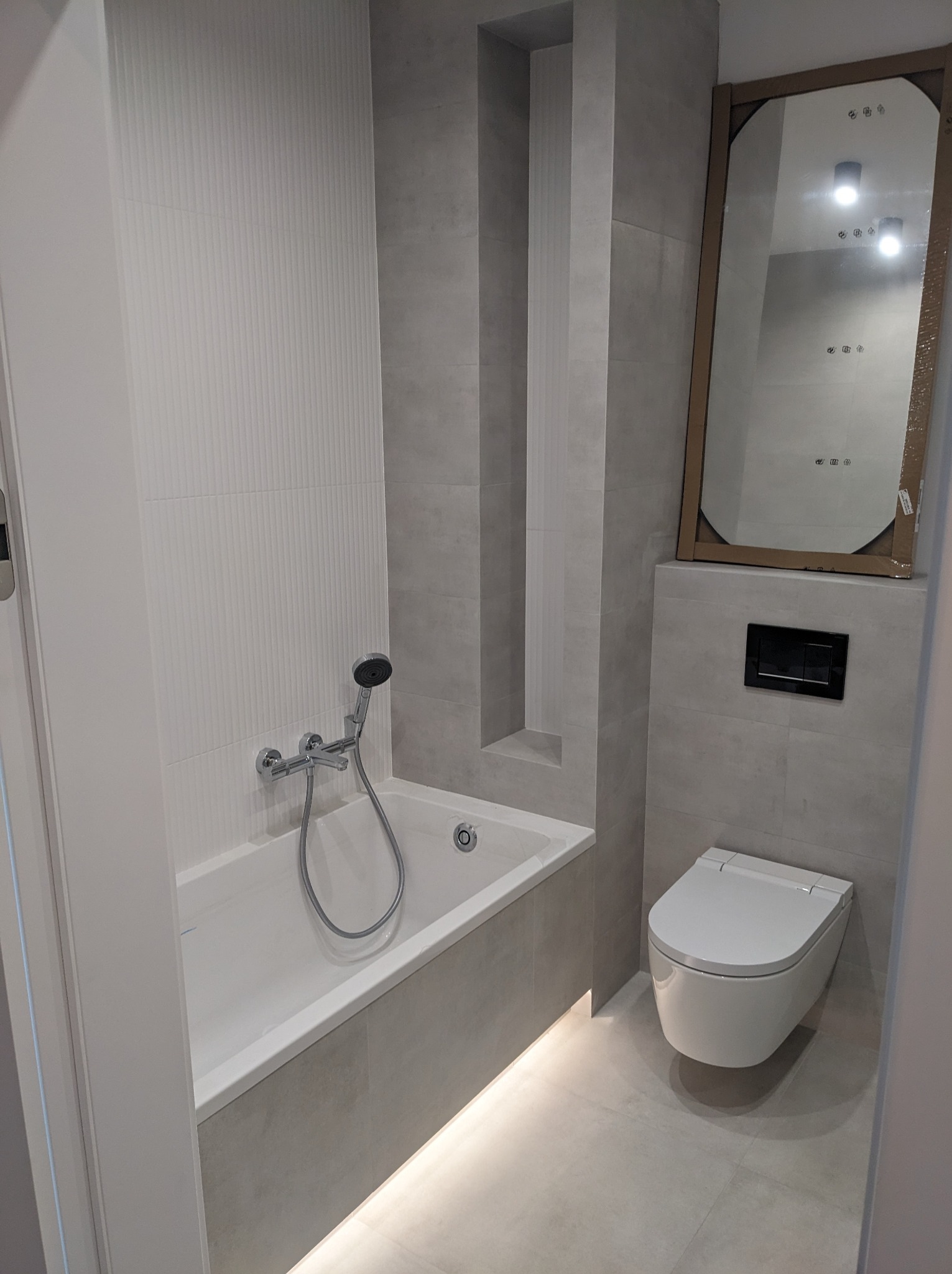 Nowoczesna łazienka w odcieniach szarości i bieli z wanną, chromowaną armaturą, podświetleniem LED u dołu wanny, wiszącą toaletą i dużym lustrem w kartonowej ramie czekającym na montaż.