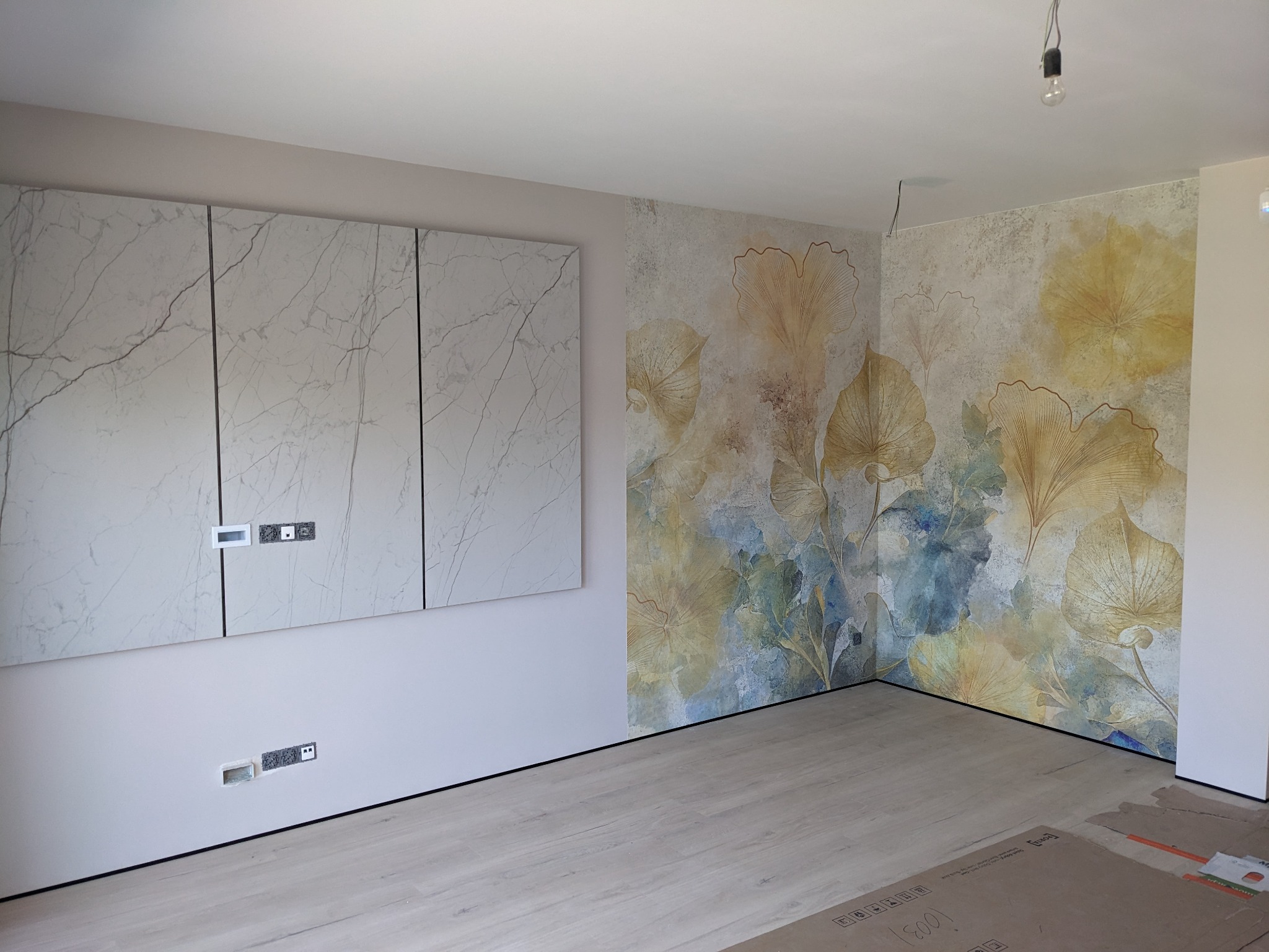 Pokój po malowaniu ścian z motywem liści ginkgo, z zamontowanymi szafkami z marmurowym wzorem i odkrytymi gniazdkami elektrycznymi, podłoga w jasnym odcieniu drewna.