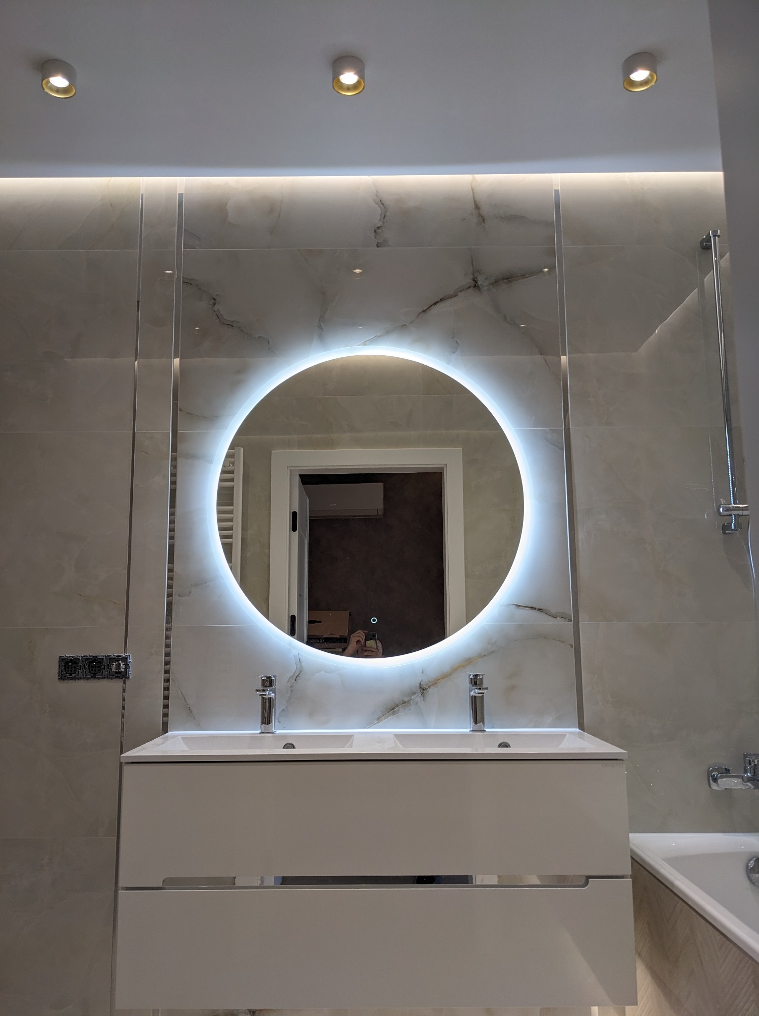 Nowoczesna łazienka z podświetlanym okrągłym lustrem, białą szafką z umywalką i marmurowymi płytkami na ścianach. W tle widoczny fragment grzejnika i drzwi do innego pomieszczenia.