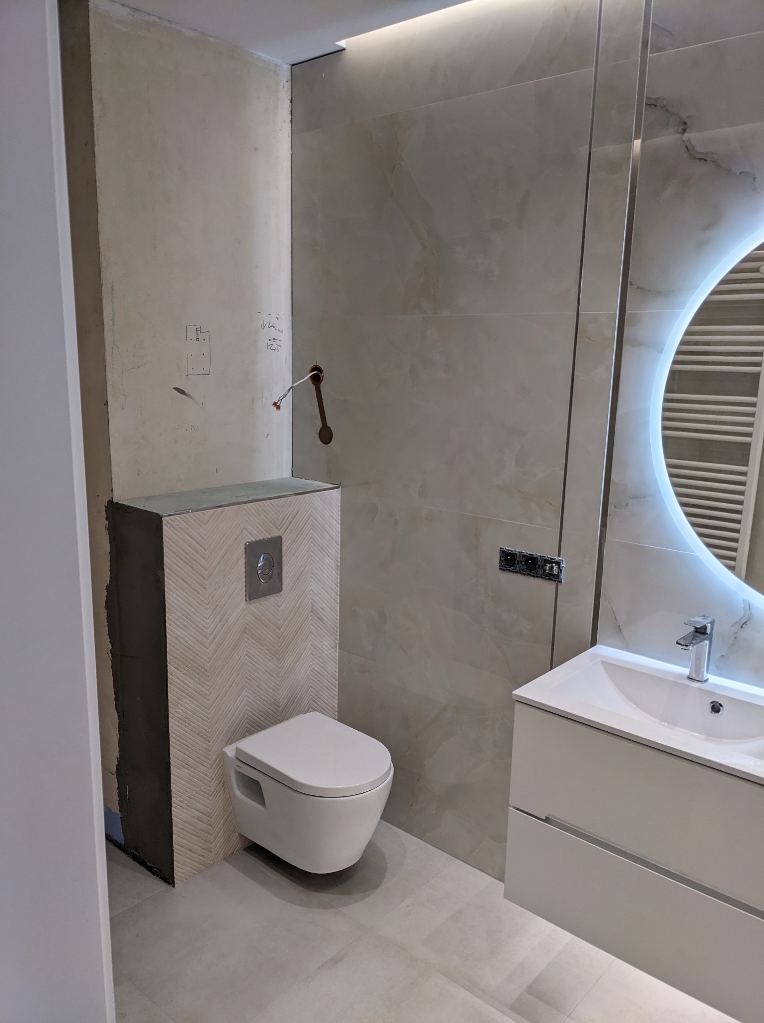 Montaż podwieszanej toalety w łazience z częściowo wykończonymi ścianami, widoczne płytki imitujące marmur, podświetlane lustro i niedokończona instalacja elektryczna.