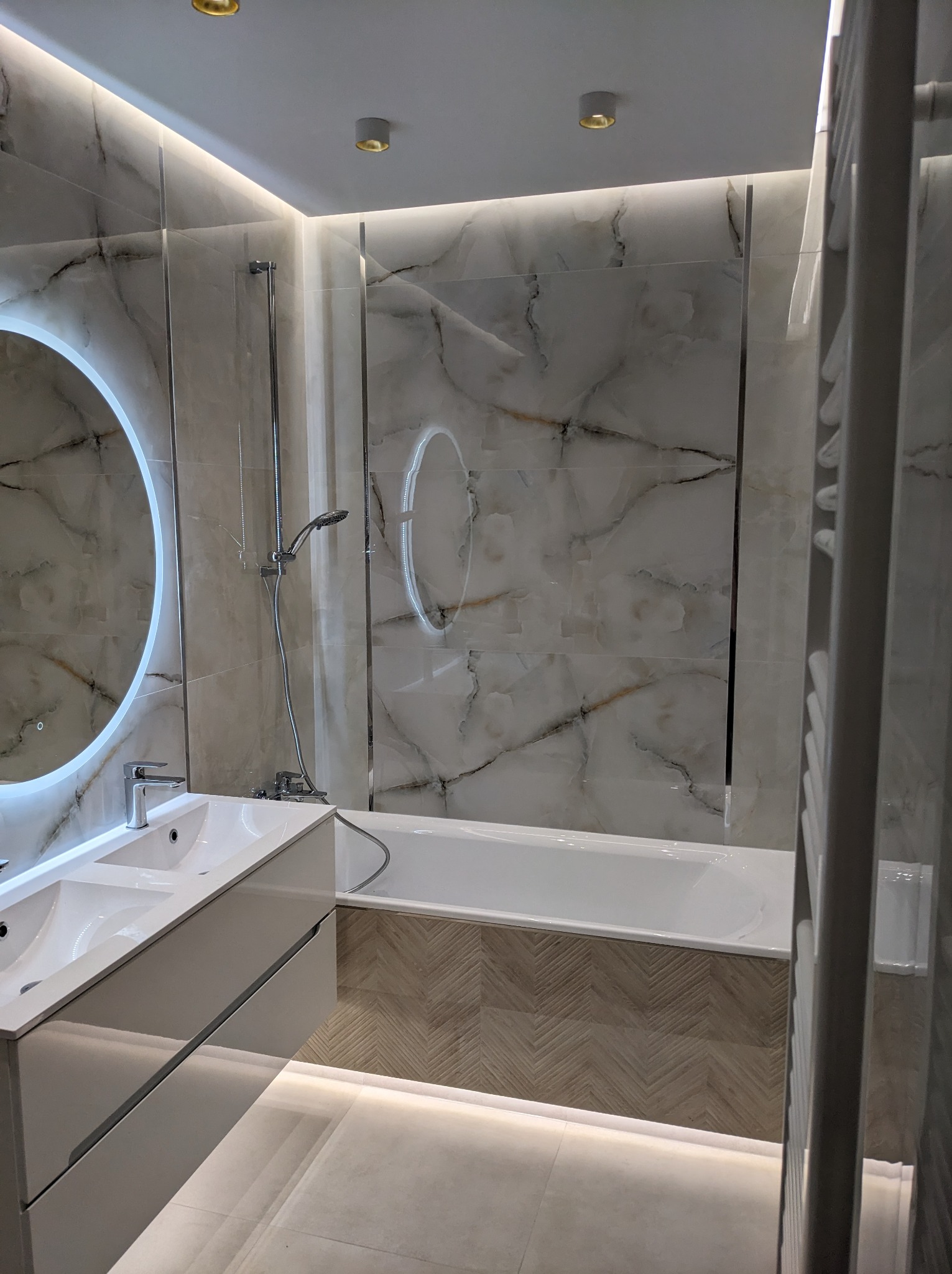 Nowoczesna łazienka w Bydgoszczy z marmurowymi płytkami, podświetlanym lustrem, białą umywalką z szafką i wanną. Podłoga wyłożona płytkami imitującymi drewno w jodełkę.