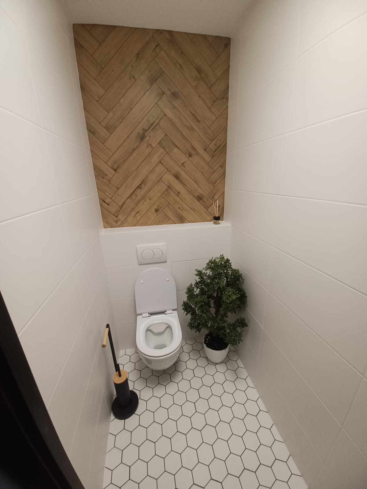 Nowoczesna toaleta z białymi płytkami, heksagonalną podłogą i drewnianym akcentem na ścianie, uzupełniona o dekoracyjną roślinę w doniczce i czarny stojak na papier toaletowy.