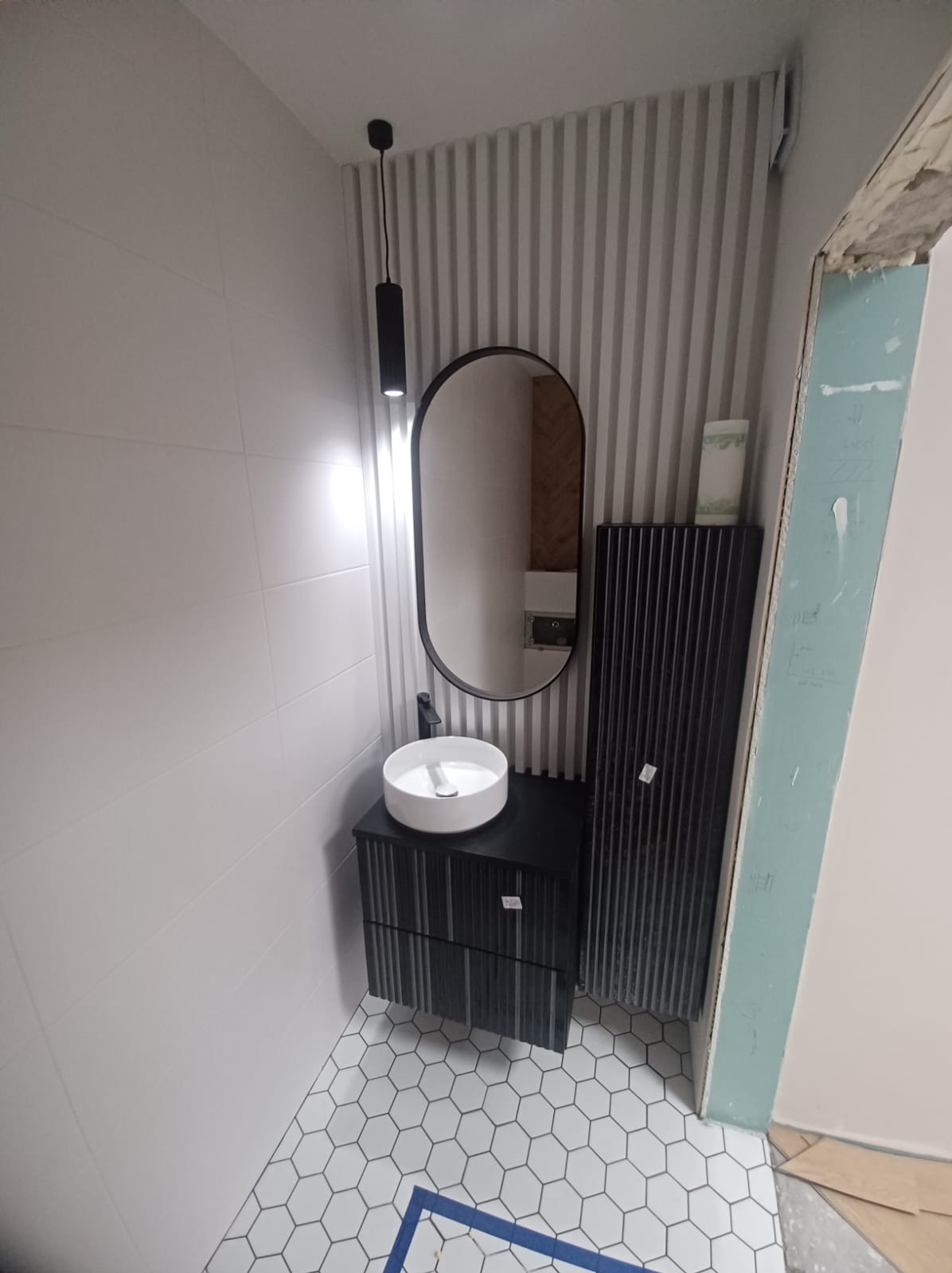 Nowoczesna łazienka z białą umywalką nablatową na czarnej szafce z frezowanymi frontami, heksagonalne płytki podłogowe z niebieskim akcentem, owalne lustro w czarnej ramie i pionowe panele ścienne...