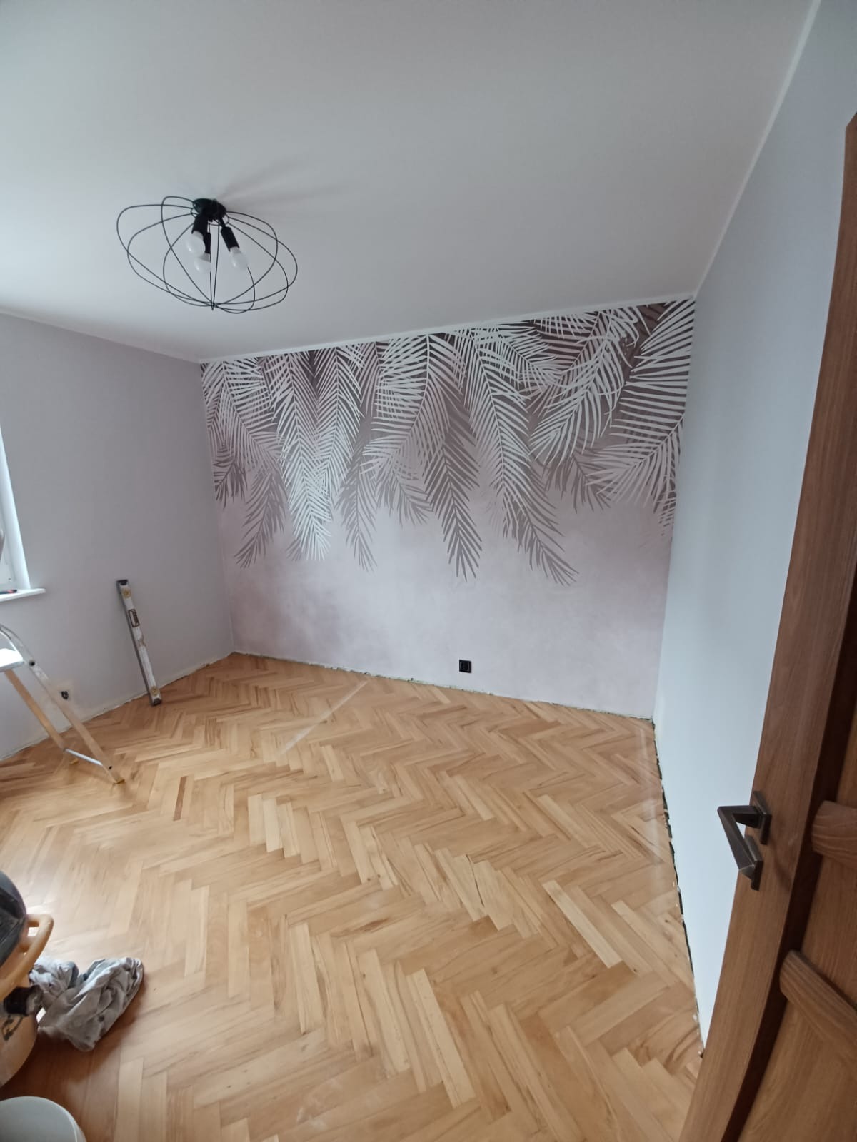 Pokój po odświeżeniu: parkiet w jodełkę, ściana z motywem liści palmowych, nowoczesna lampa sufitowa, drabina oparta o ścianę.