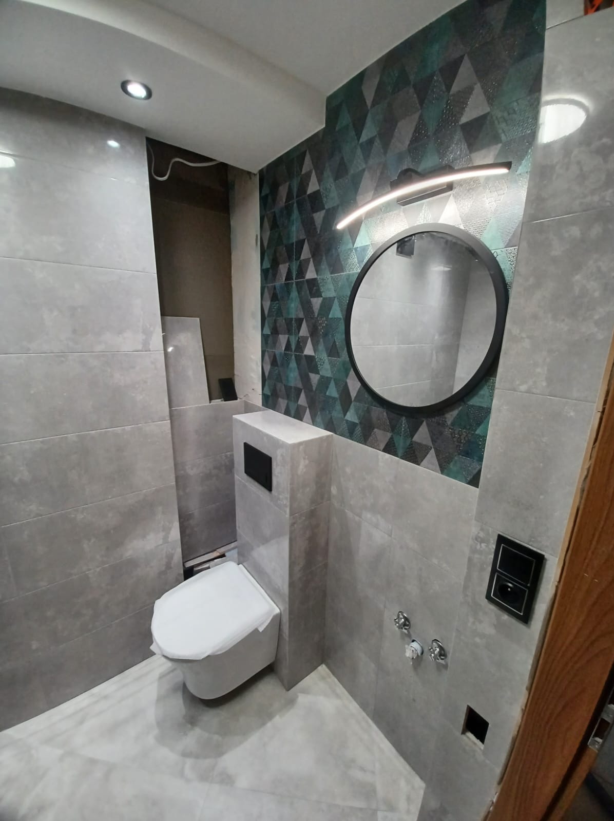 Nowoczesna łazienka z szarymi płytkami imitującymi beton, akcentem w postaci ściany z geometrycznym wzorem w odcieniach zieleni i szarości, okrągłym lustrem z podświetleniem LED oraz podwieszaną...