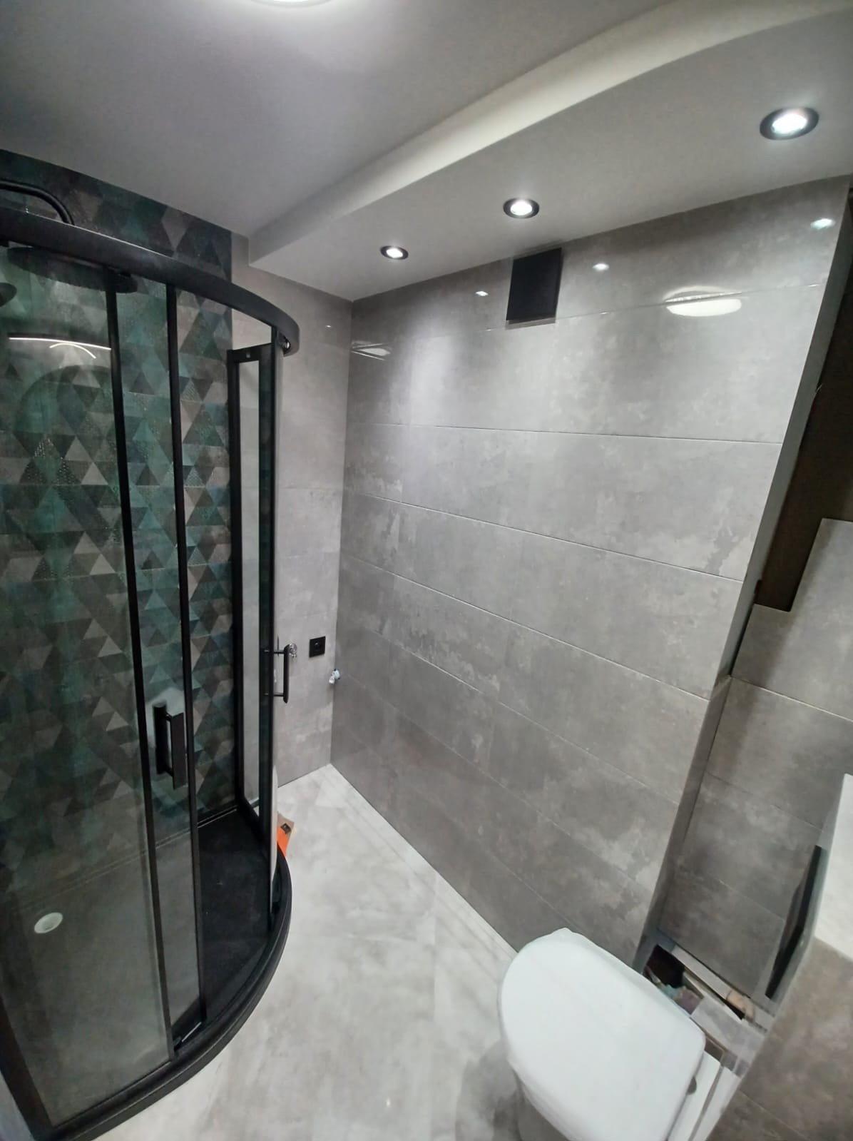 Wykończona łazienka z kabiną prysznicową z czarnymi profilami i geometrycznym wzorem na szkle, szare płytki ścienne ułożone poziomo, biała toaleta, i punktowe oświetlenie sufitowe.