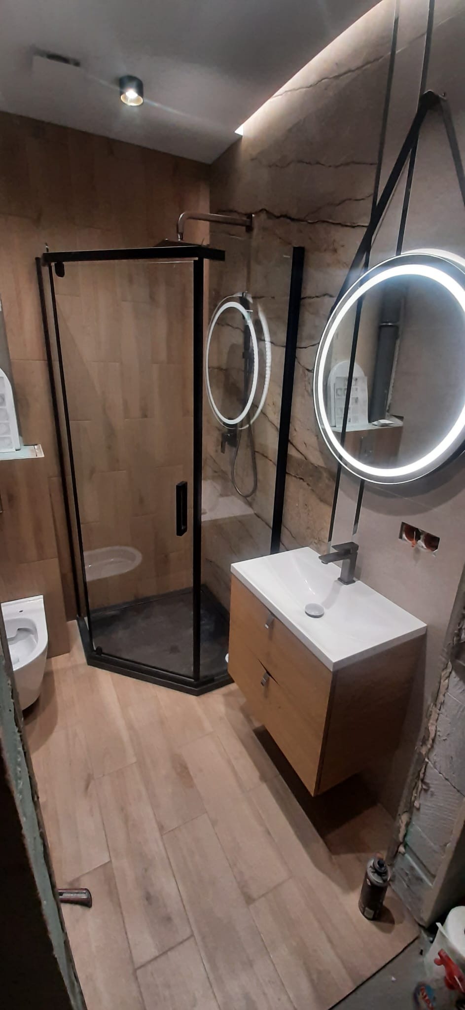 Nowoczesna łazienka po remoncie: kabina prysznicowa z czarnymi profilami, okrągłe lustra z podświetleniem LED, drewniana szafka z umywalką, płytki imitujące drewno na podłodze i ścianie, fragment...