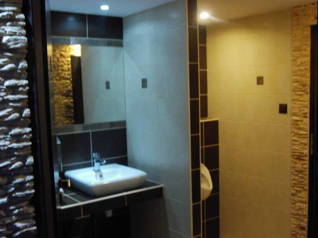 Nowoczesna łazienka z umywalką w kształcie kwadratu, lustrem z czarną ramą, oraz ścianą wyłożoną płytkami imitującymi kamień, obok znajduje się pisuar i ściana z jasnymi płytkami.