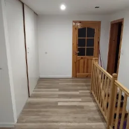 Korytarz z drewnianą balustradą, panelami podłogowymi imitującymi drewno, białą szafą przesuwną i drewnianymi drzwiami z szybkami w tle.