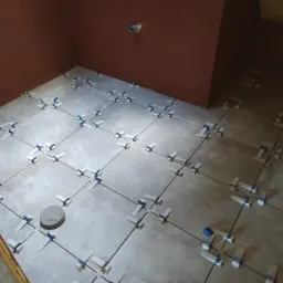 Szare płytki podłogowe w trakcie układania, z użyciem białych krzyżyków i niebieskich klinów poziomujących, na tle pomarańczowych ścian w surowym stanie.