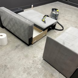 Pranie Tapicerek meblowych oraz samochodowych - Rozłożona szara kanapa z wysuniętą szufladą, częściowo wyczyszczona, obok stoi odkurzacz piorący i rolka papieru.
