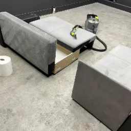 Rozłożona szara kanapa z wysuniętą szufladą, częściowo wyczyszczona, obok stoi odkurzacz piorący i rolka papieru.