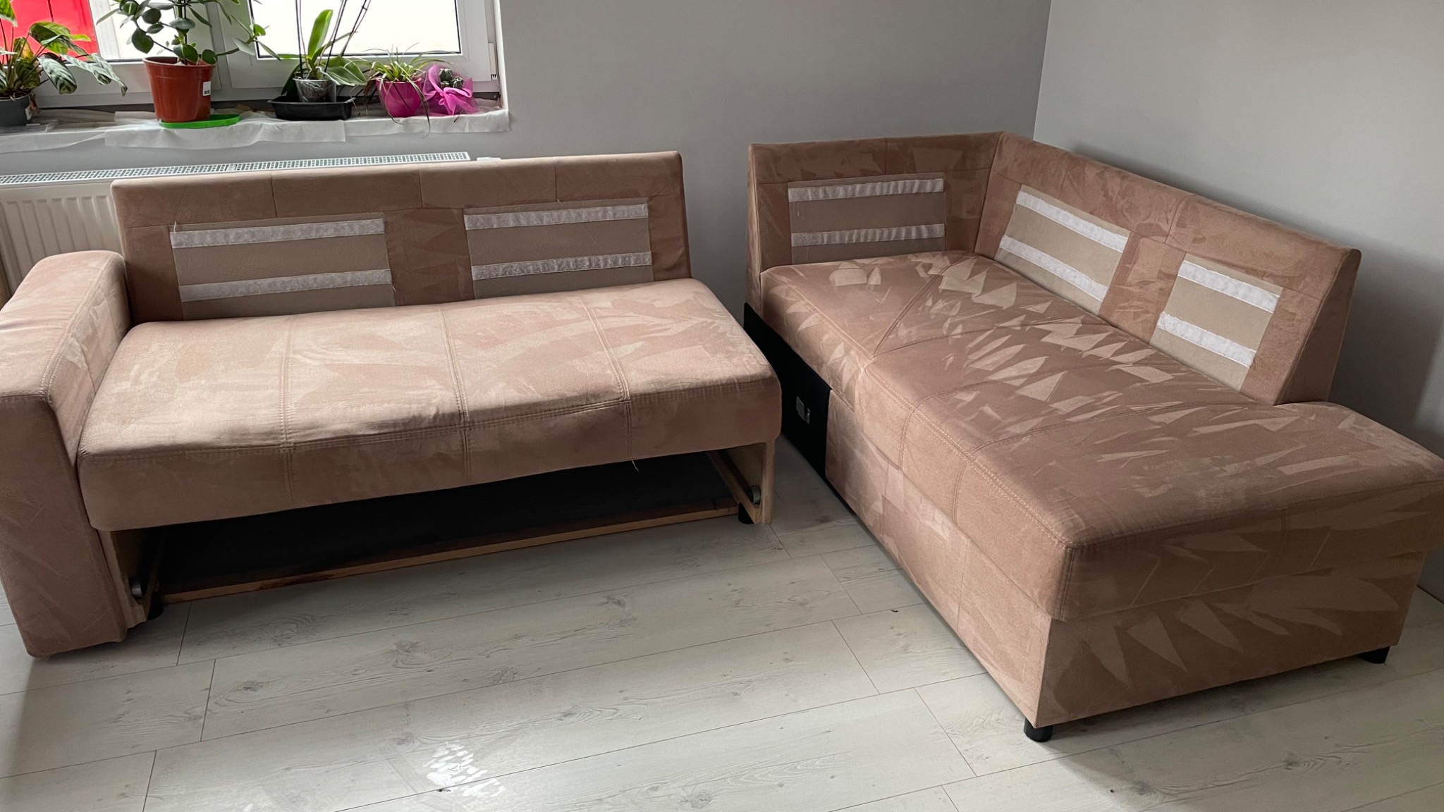 Rozłożona narożna kanapa w odcieniach beżu z widocznym mechanizmem wysuwania, ustawiona na jasnej podłodze, z roślinami w tle na parapecie okiennym.