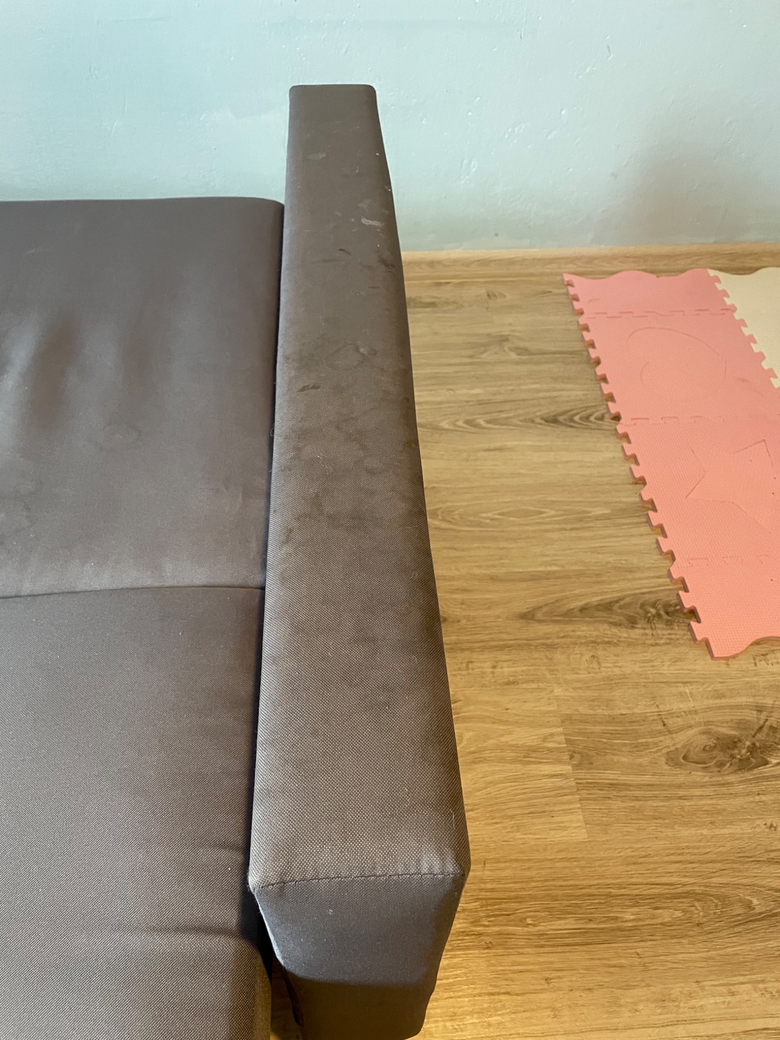 Szary narożnik z widocznymi plamami na tapicerce, fragment podłogi z panelami i różowo-beżowymi puzzlami piankowymi w tle.