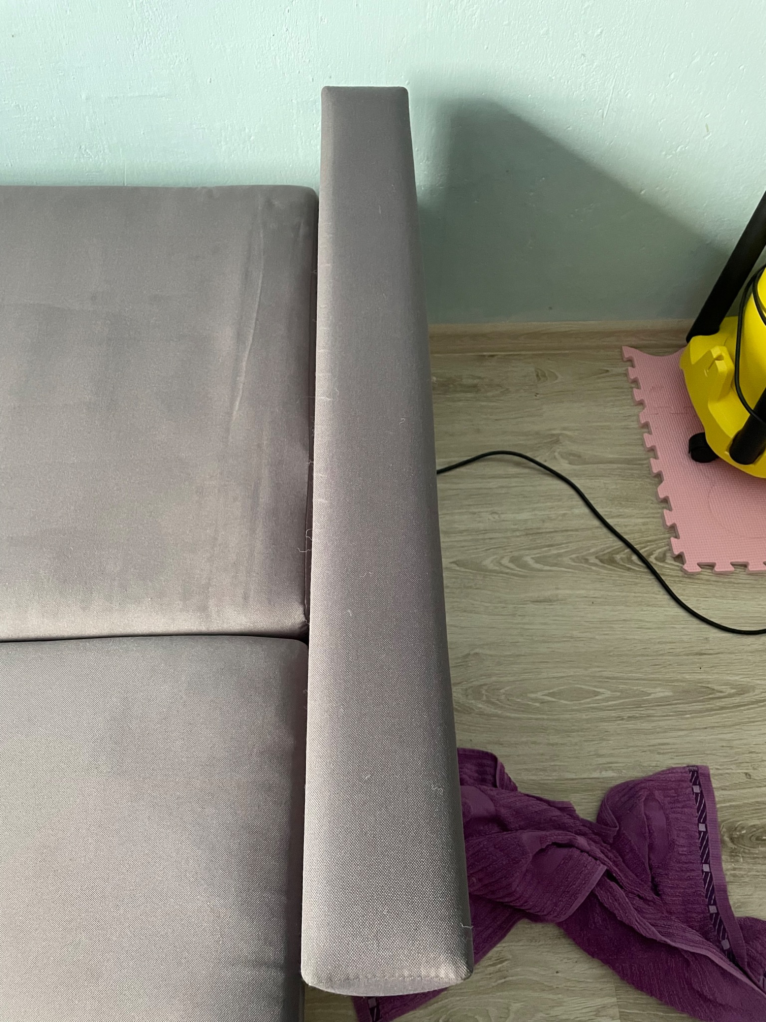 Fragment szarej kanapy z widocznym bokiem i siedziskiem, obok leży fioletowy ręcznik, a w tle żółty odkurzacz piorący.
