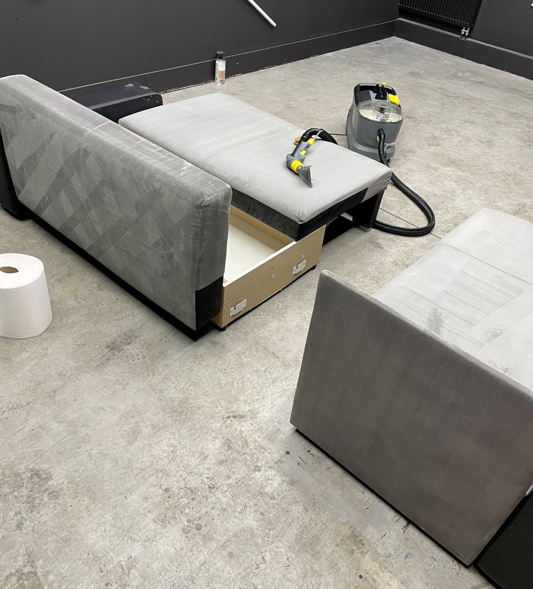 Rozłożona szara kanapa z wysuniętą szufladą, częściowo wyczyszczona, obok stoi odkurzacz piorący i rolka papieru.