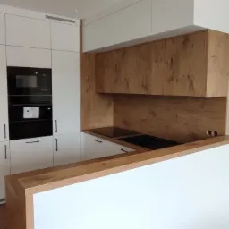 Nowoczesna kuchnia z białymi szafkami i drewnopodobnymi akcentami, wbudowany piekarnik i płyta grzewcza, widoczna drabina malarska w tle.