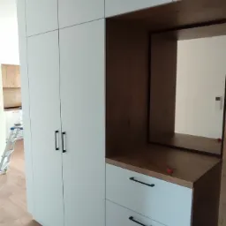 Biała szafa z zabudową i trzema szufladami z czarnymi uchwytami, z drewnianym blatem i zabudowanym lustrem, widoczna z boku na tle drewnianej podłogi.