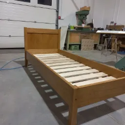 Drewniane łóżko jednoosobowe z jasnego drewna w warsztacie stolarskim, widoczne lamele pod materac, w tle maszyny stolarskie i brama garażowa.