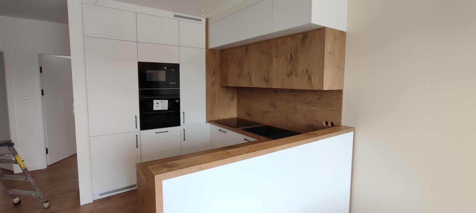 Nowoczesna kuchnia z białymi szafkami i drewnopodobnymi akcentami, wbudowany piekarnik i płyta grzewcza, widoczna drabina malarska w tle.