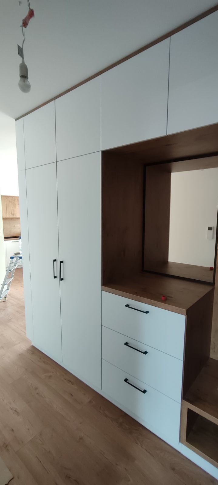 Biała szafa z zabudową i trzema szufladami z czarnymi uchwytami, z drewnianym blatem i zabudowanym lustrem, widoczna z boku na tle drewnianej podłogi.
