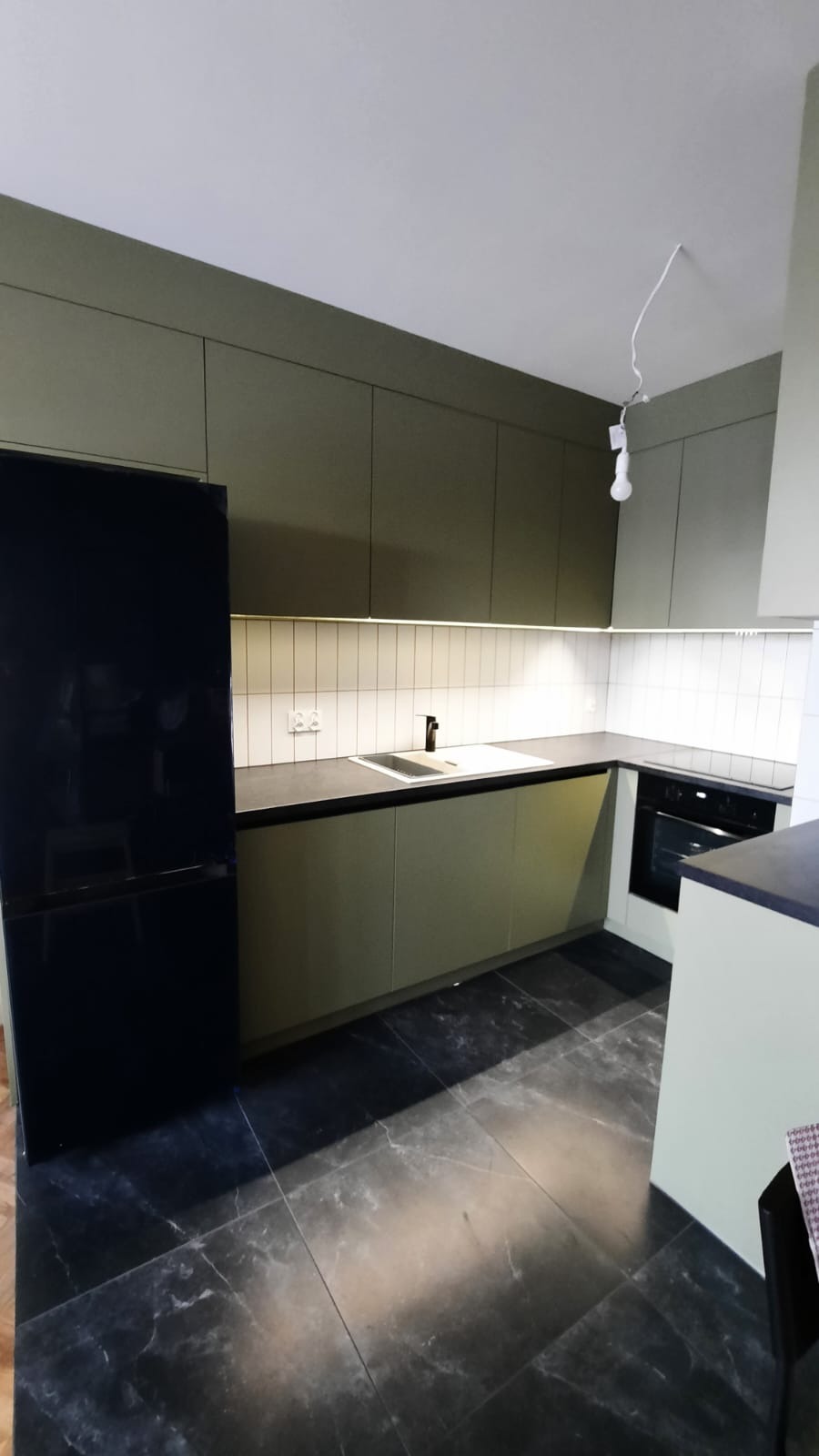 Nowoczesna kuchnia w odcieniach oliwkowej zieleni, z czarnym blatem i marmurową podłogą. Białe, wąskie kafelki nad blatem, oświetlenie LED pod szafkami. Czarna lodówka side-by-side po lewej stronie...