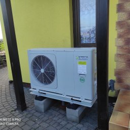 Centauron Sp. z o.o. - Zewnętrzna jednostka pompy ciepła Beretta na betonowych blokach przed żółtą ścianą domu z oknem i ozdobną elewacją. Pies w tle.