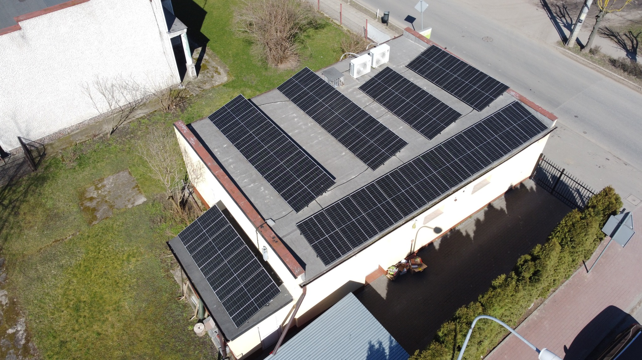 Widok z góry na budynek z panelami fotowoltaicznymi na dachu. Dodatkowo na dachu widoczne jednostki klimatyzacji. Obok budynku drzewa i droga.