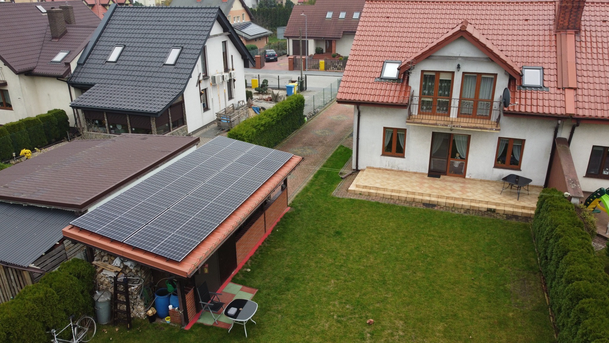 Widok z góry na dom z panelami słonecznymi na dachu wiaty, zielony trawnik i zabudowę jednorodzinną w tle. Perspektywa z lotu ptaka.
