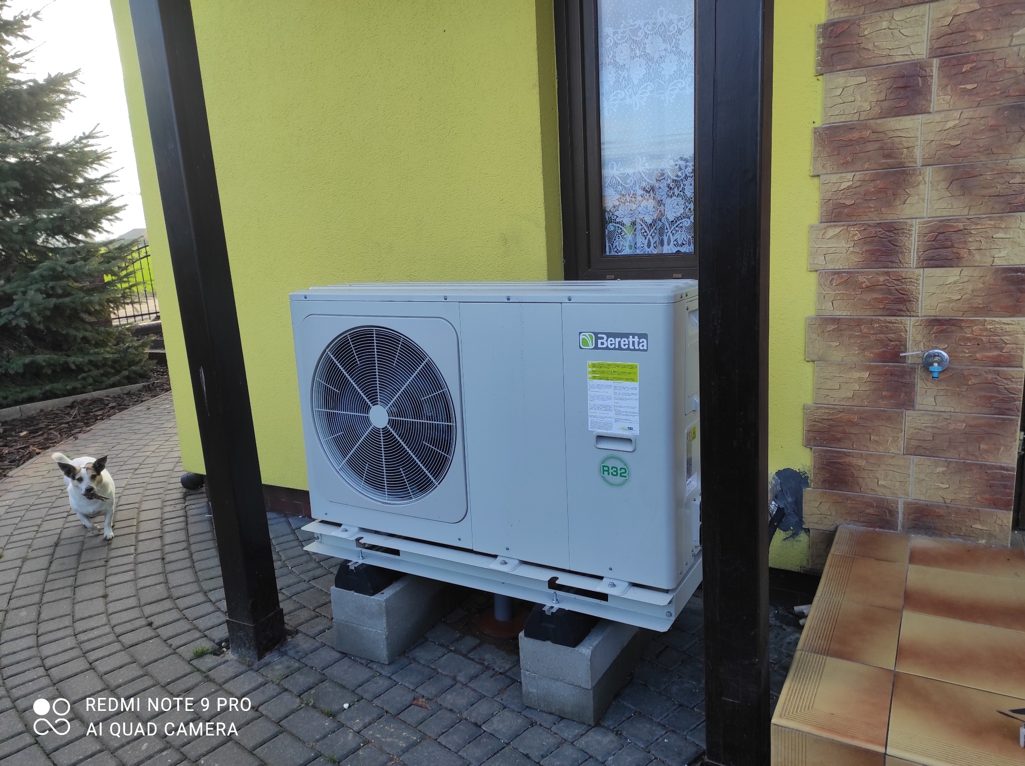 Zewnętrzna jednostka pompy ciepła Beretta na betonowych blokach przed żółtą ścianą domu z oknem i ozdobną elewacją. Pies w tle.