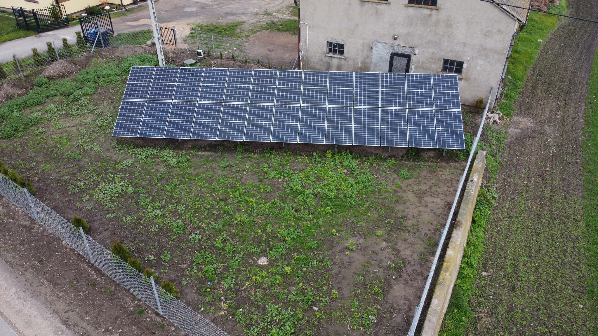 Widok z góry na panele fotowoltaiczne zamontowane na gruncie obok budynku. Panele ułożone w rzędach, otoczone zielenią i ogrodzeniem. Ujęcie z drona.