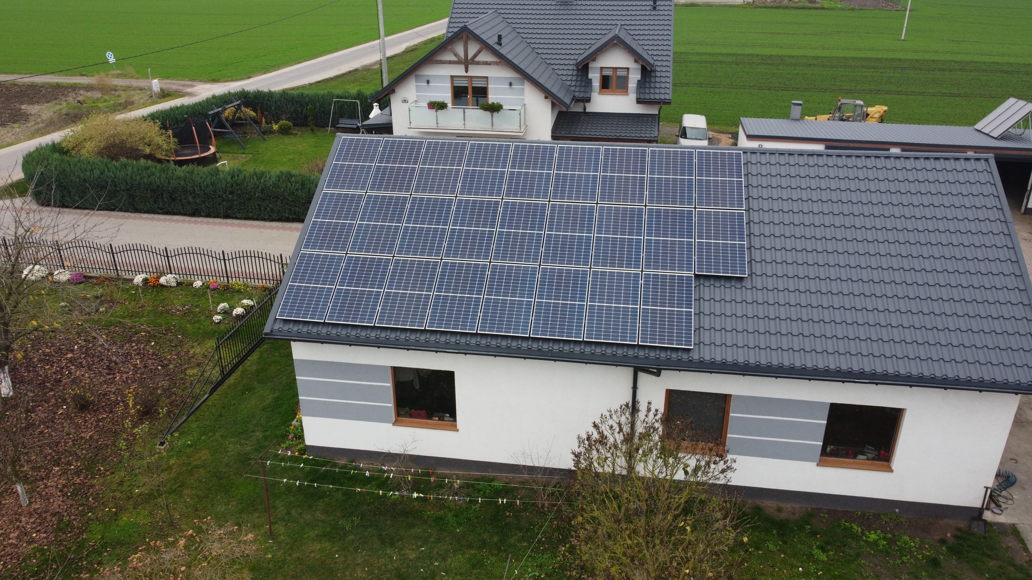 Instalacja fotowoltaiczna na dachu domu jednorodzinnego z szarej blachodachówki, widok z góry na zielone otoczenie i drugi budynek w tle.