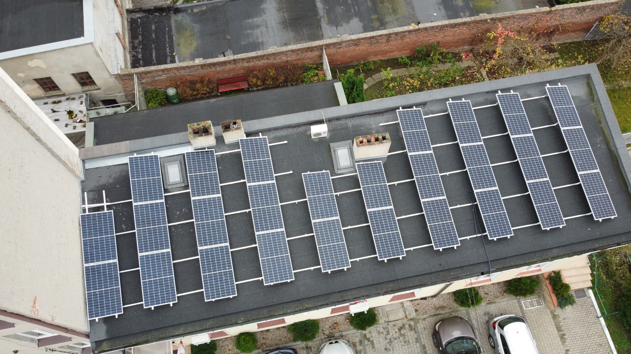 Widok z góry na rząd paneli fotowoltaicznych na dachu budynku z kominami i oknami dachowymi, otoczonego zielenią i budynkami mieszkalnymi.