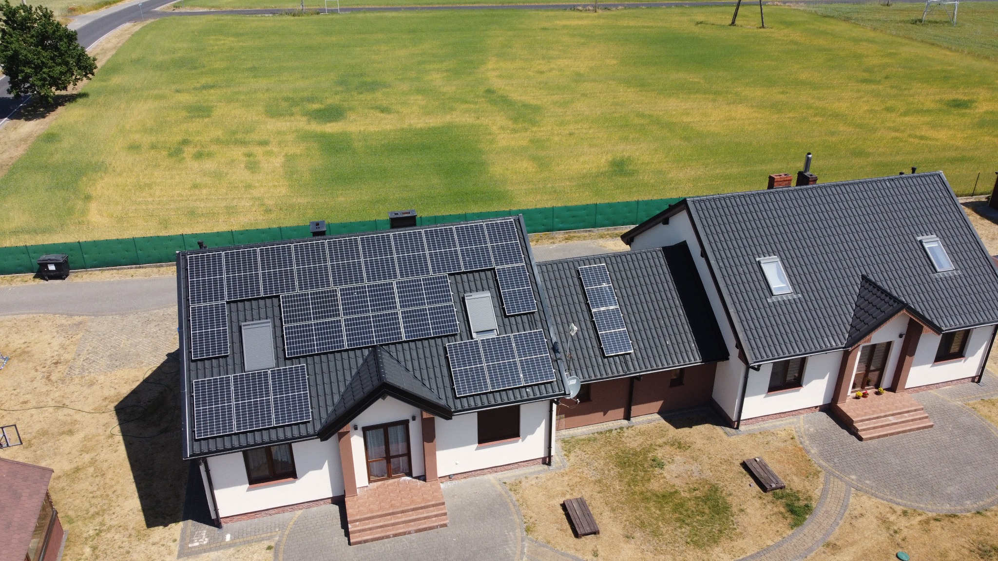Widok z góry na dom z panelami fotowoltaicznymi na dachu. Panele zamontowane na szarym dachu z dachówki. W tle zielone pole i droga.