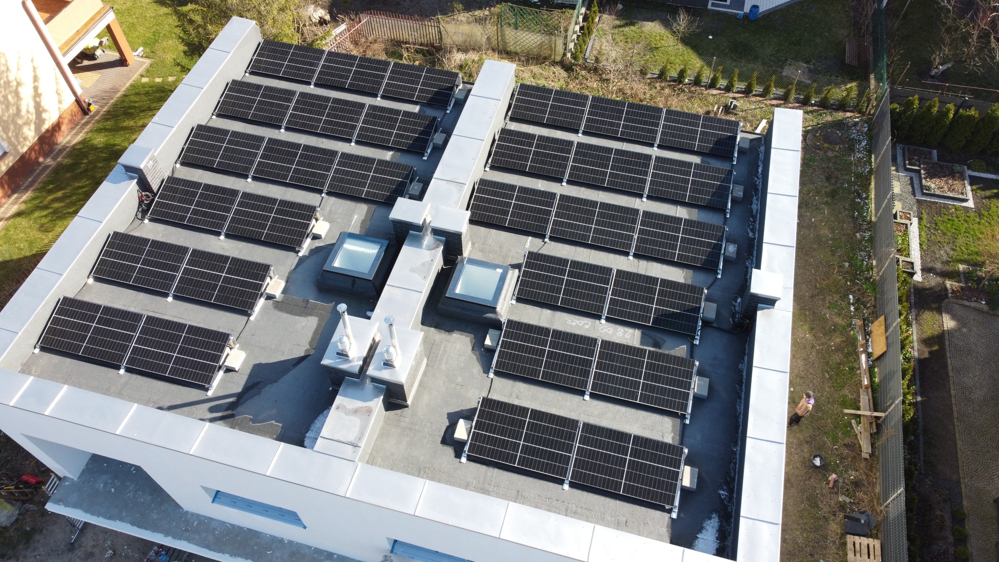 Widok z góry na płaski dach z panelami słonecznymi, dwa świetliki i elementy wentylacji. W tle zieleń, częściowo ocieniona.