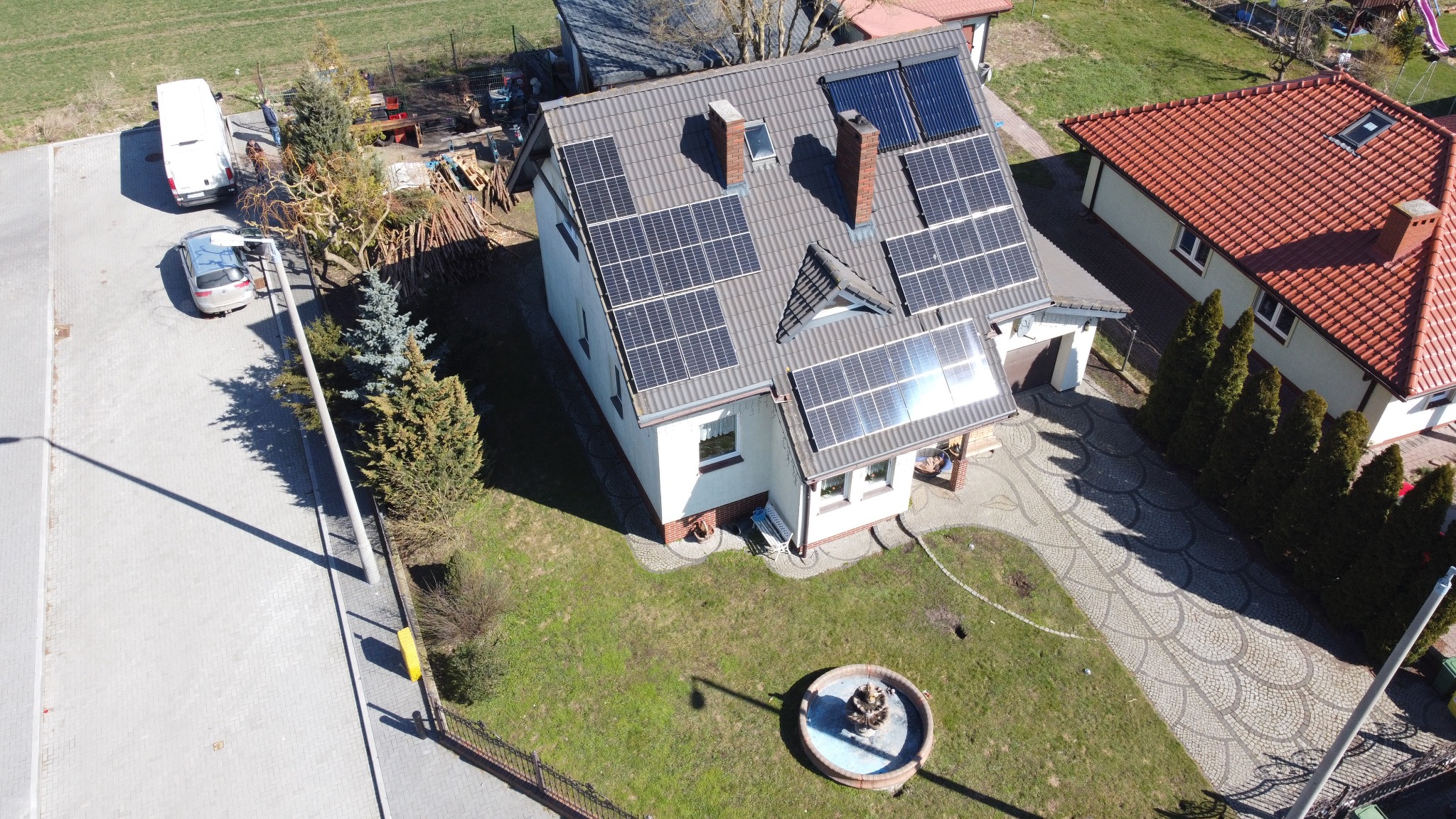 Dom jednorodzinny z panelami fotowoltaicznymi i kolektorami słonecznymi na dachu, widziany z lotu ptaka. Na posesji fontanna i podjazd z kostki.