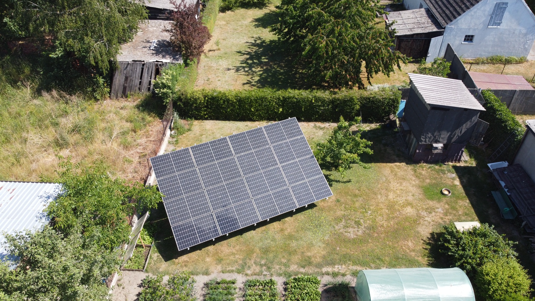 Widok z góry na panele fotowoltaiczne na trawniku, obok budynki gospodarcze i szklarnia. Panele ułożone w prostokąt, otoczone zielenią i zabudową.