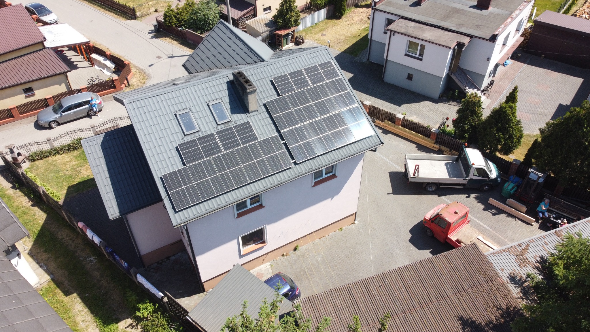 Dom z szarym dachem i panelami fotowoltaicznymi, widok z góry. Na podwórzu zaparkowane dwa samochody dostawcze i osobowy. Częściowo widoczna ulica i sąsiednie budynki.