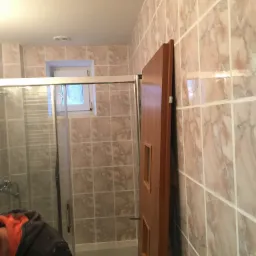 Wykończona łazienka z kabiną prysznicową, brązowymi płytkami imitującymi marmur, drewnianymi drzwiami z małymi okienkami i małym oknem nad kabiną.