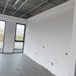 Wnętrze biura w stanie deweloperskim z surowym betonowym stropem podwieszonym metalową konstrukcją, białymi ścianami z widocznymi instalacjami elektrycznymi i hydraulicznymi, szarą podłogą z płytek...