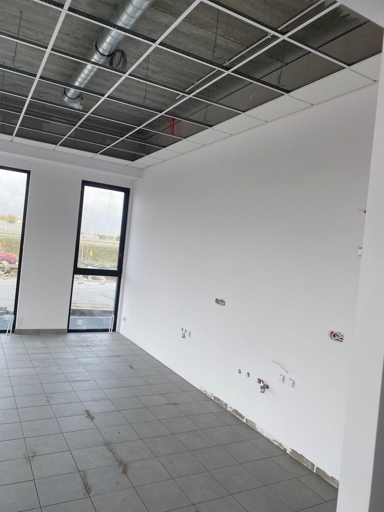 Wnętrze biura w stanie deweloperskim z surowym betonowym stropem podwieszonym metalową konstrukcją, białymi ścianami z widocznymi instalacjami elektrycznymi i hydraulicznymi, szarą podłogą z płytek...