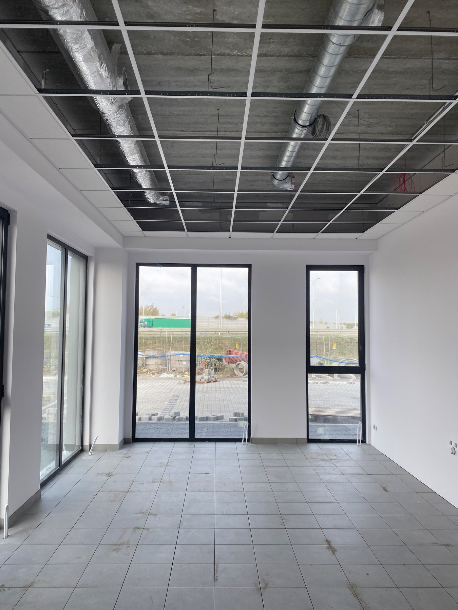 Wnętrze biura w stanie deweloperskim z widoczną instalacją wentylacyjną pod sufitem, surowym betonem i rozłożoną siatką pod płyty kartonowo-gipsowe, duże okna z widokiem na plac budowy.