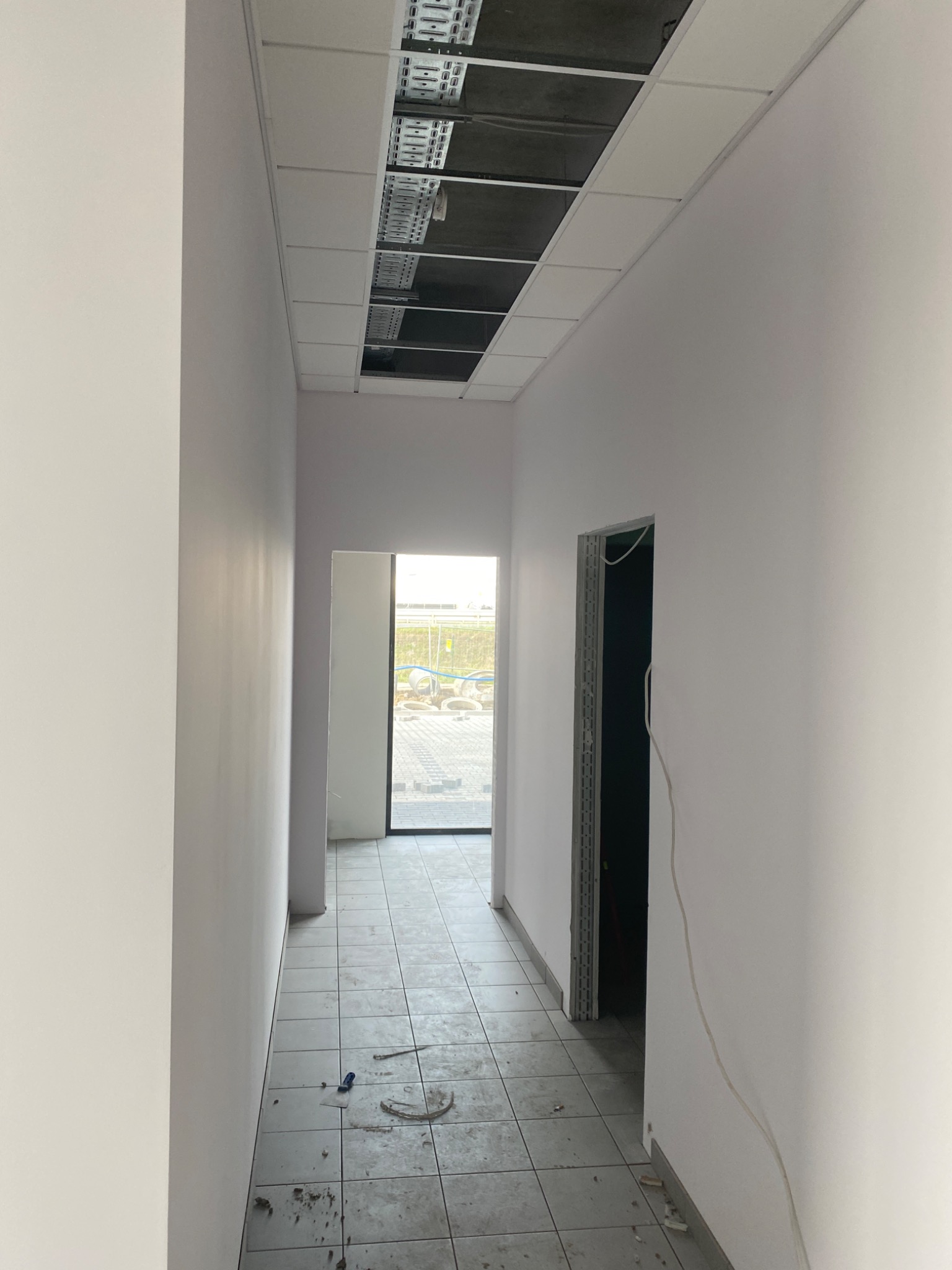 Korytarz w trakcie wykańczania, widoczne białe ściany, szara podłoga z płytek, sufit z otwartą instalacją i otworem, oraz ościeżnice drzwi bez drzwi, na podłodze leżą drobne narzędzia.