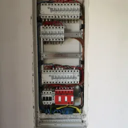 Rozdzielnica elektryczna z zabezpieczeniami przepięciowymi zamontowana w ścianie, widoczne przewody w kolorach niebieskim, żółtym i brązowym.