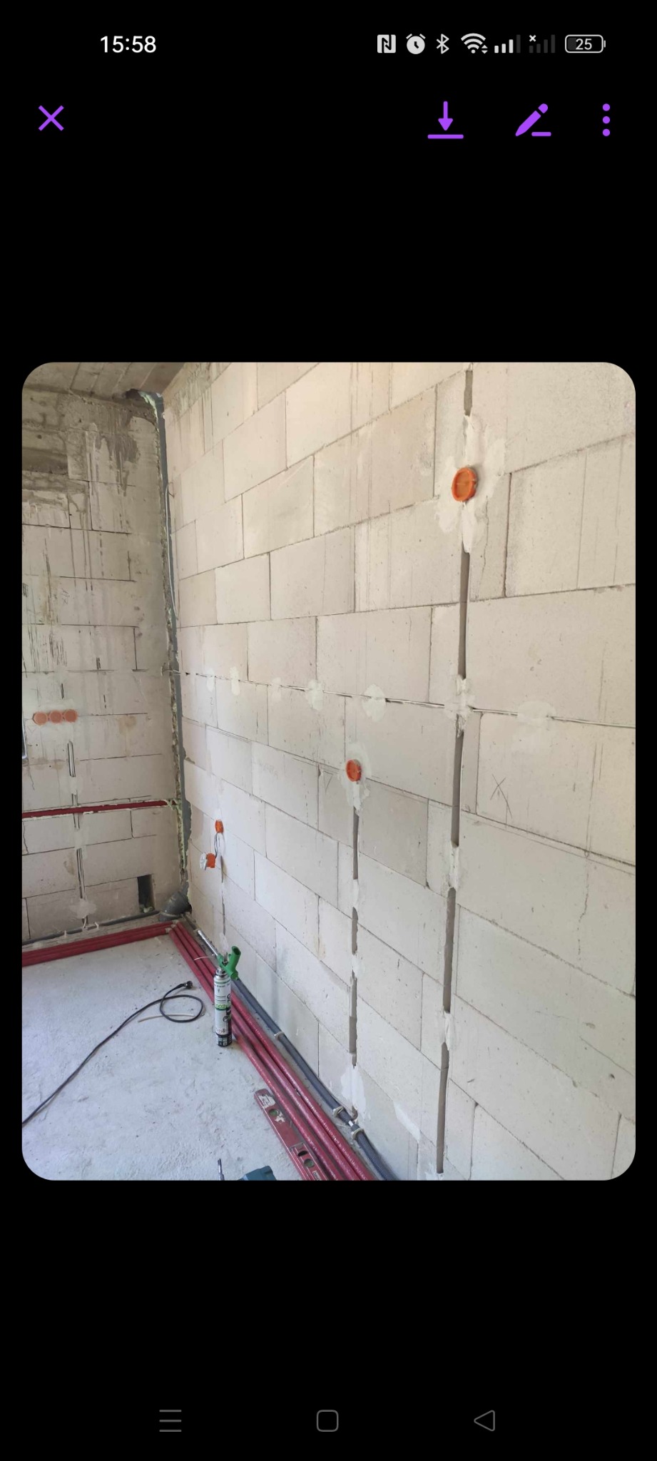 Ściana z betonu komórkowego z bruzdami wypełnionymi gipsem, pomarańczowe puszki elektryczne, czerwone rury instalacyjne, poziomica i pistolet do pianki montażowej na podłodze w surowym wnętrzu.