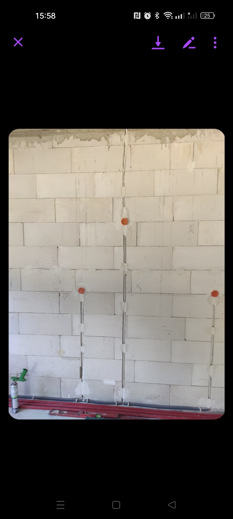 Ściana z betonu komórkowego z wyciętymi bruzdami pod przewody elektryczne, widoczne pomarańczowe puszki instalacyjne i metalowe rurki osłonowe, na dole leżą poziomice i pistolet do pianki montażowej.